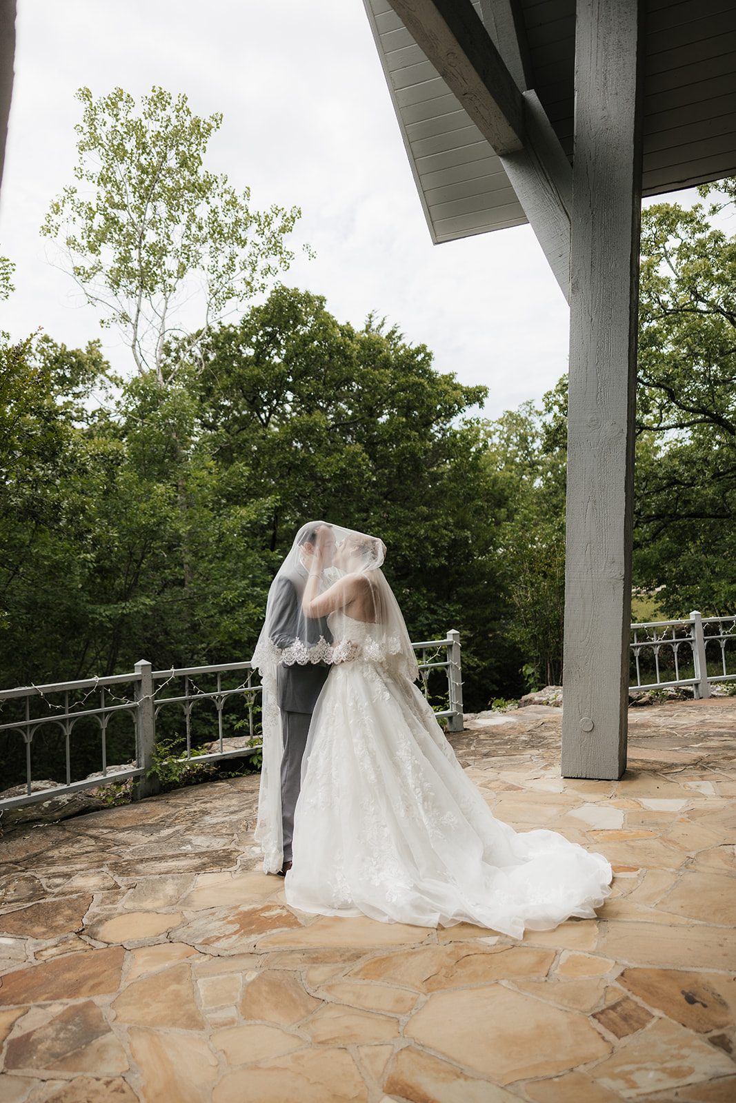 A bride and groom stand on a stone patio, sharing a kiss under the bride’s veil with trees and a metal railing in the background for a Camp Loughridge Wedding