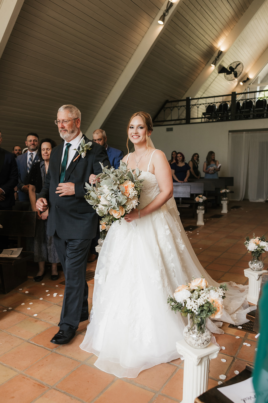 A bride and groom stand at the altar in a chapel with large windows, surrounded by their wedding party and guests seated in pews for a Camp Loughridge Wedding