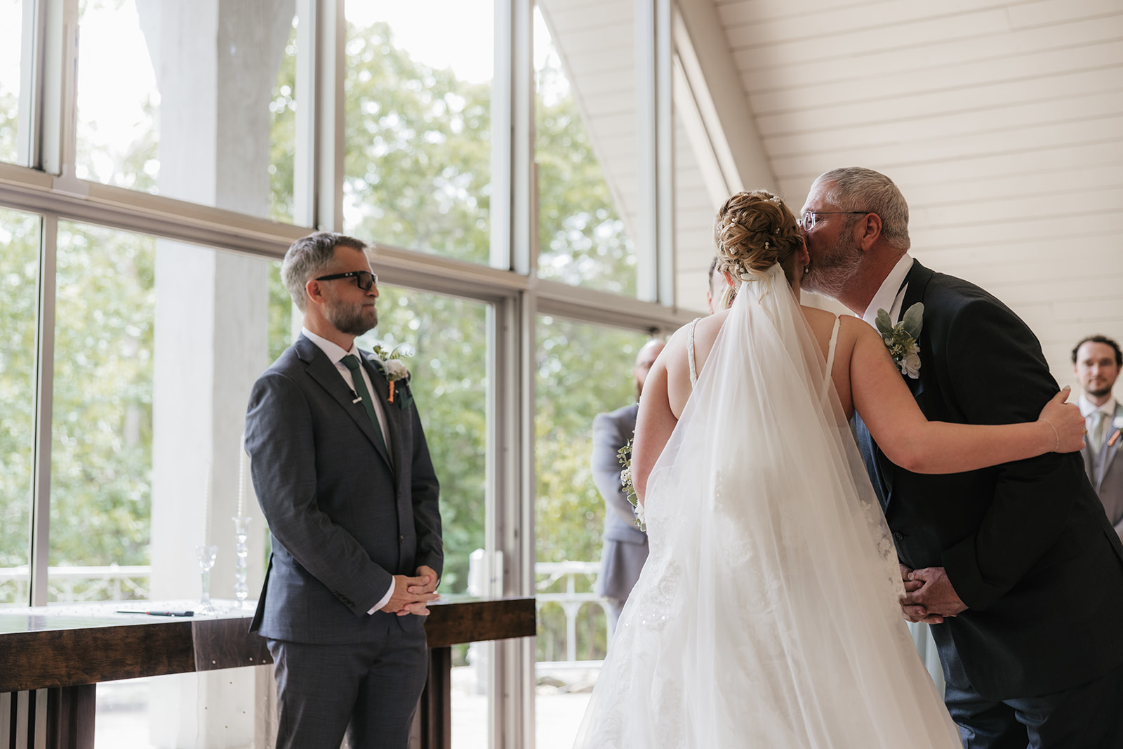 A bride and groom stand at the altar in a chapel with large windows, surrounded by their wedding party and guests seated in pews for a Camp Loughridge Wedding