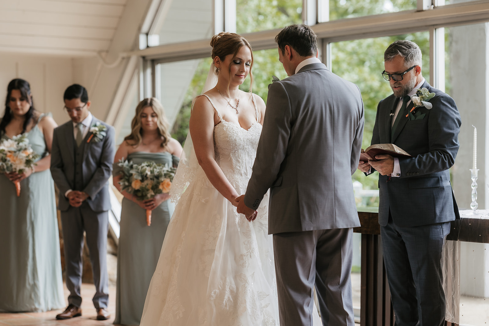 A bride and groom stand at the altar in a chapel with large windows, surrounded by their wedding party and guests seated in pews for a Camp Loughridge Wedding