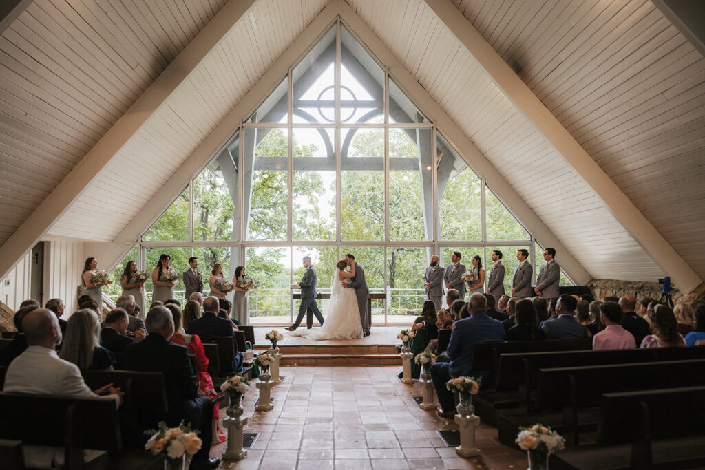 A bride and groom stand at the altar in a chapel with large windows, surrounded by their wedding party and guests seated in pews for a Camp Loughridge Wedding