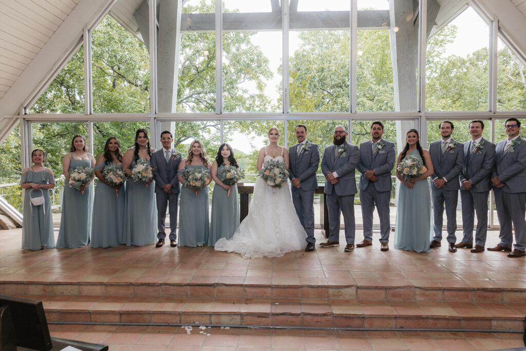 A wedding party stands in a row indoors, with the bride and groom in the center, bridesmaids and groomsmen on either side, all wearing formal attire.