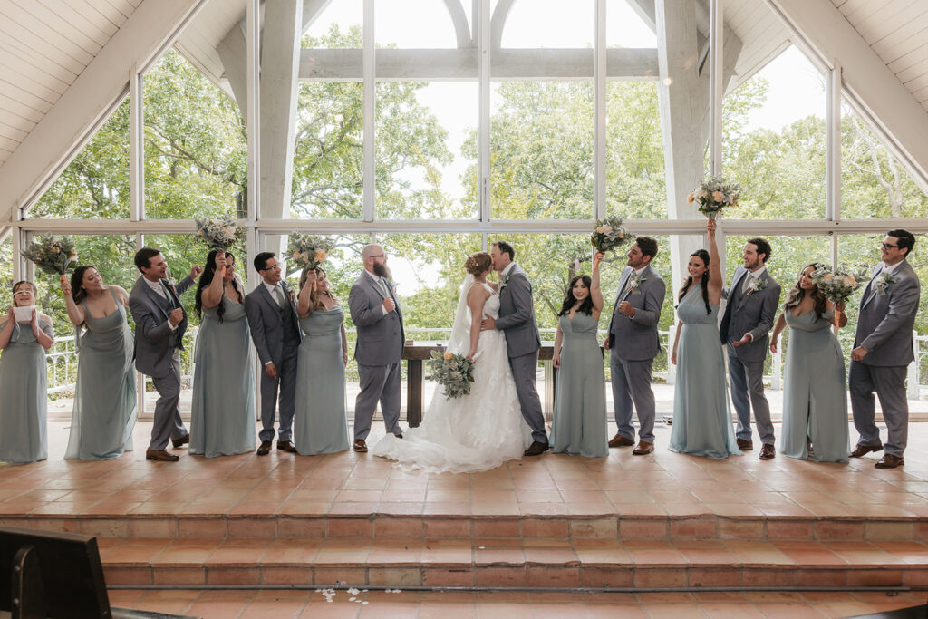 A wedding party stands in a row indoors, with the bride and groom in the center, bridesmaids and groomsmen on either side, all wearing formal attire.