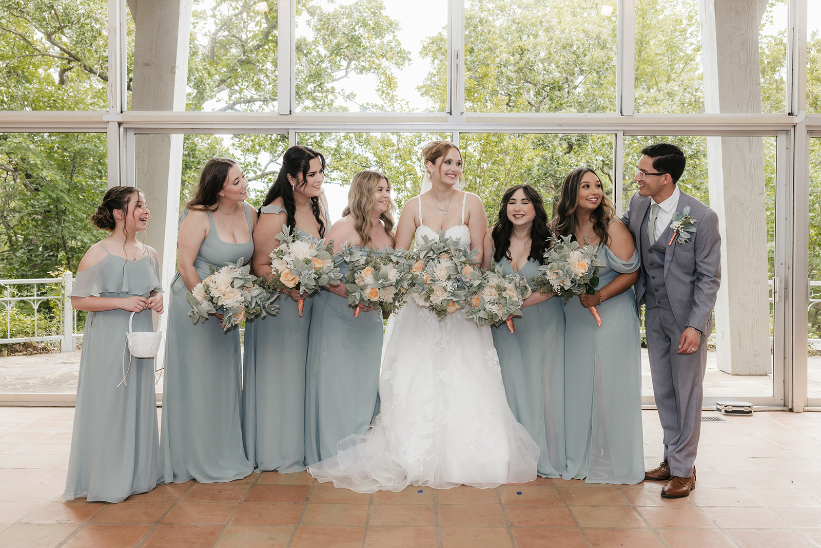A wedding party stands in a row indoors, with the bride and groom in the center, bridesmaids and groomsmen on either side, all wearing formal attire at a Camp Loughridge Wedding