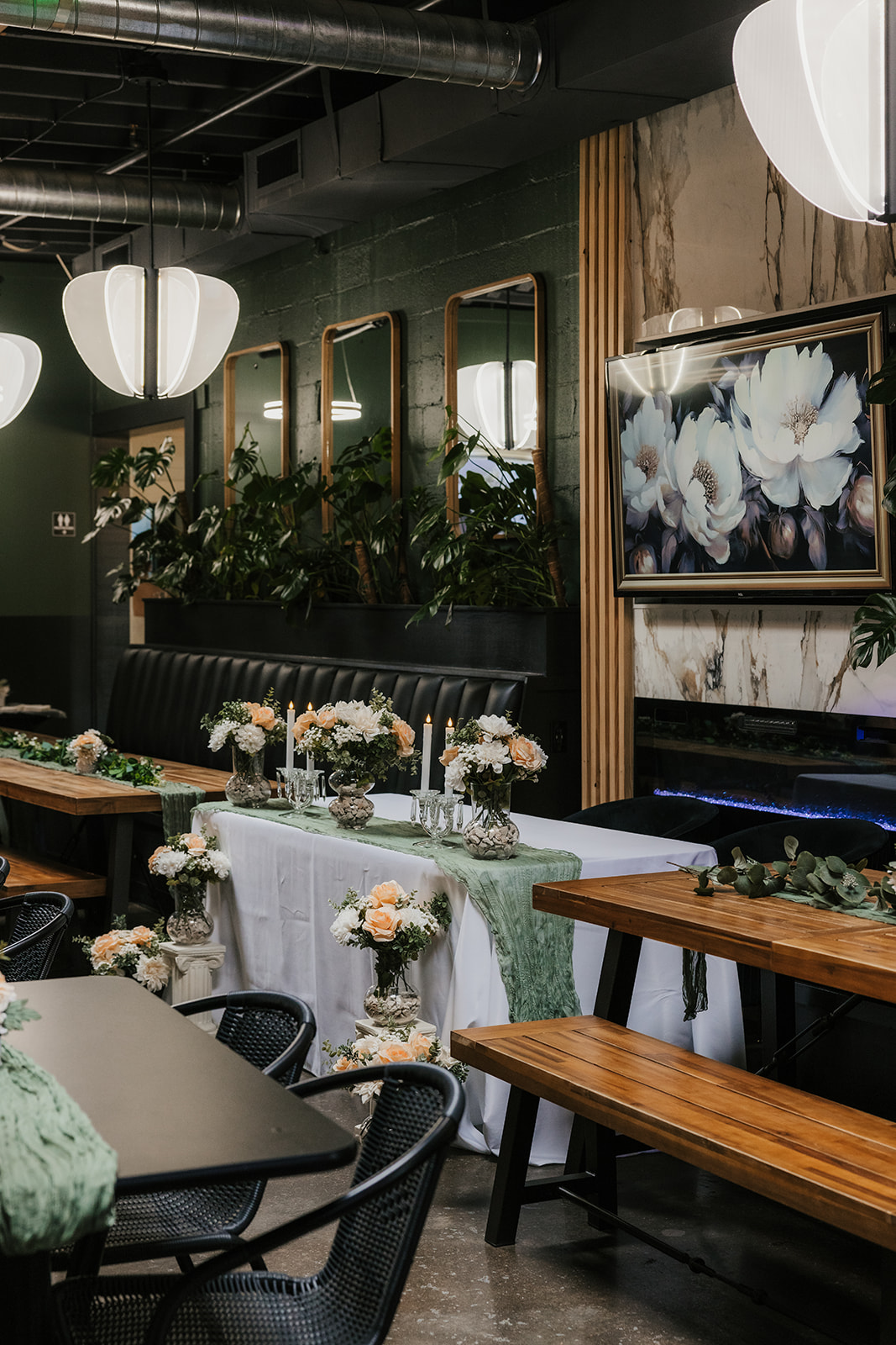 A decorated dining area with long wooden tables, floral centerpieces, green table runners, wall mirrors, and large flower artwork on the wall at a Camp Loughridge Wedding