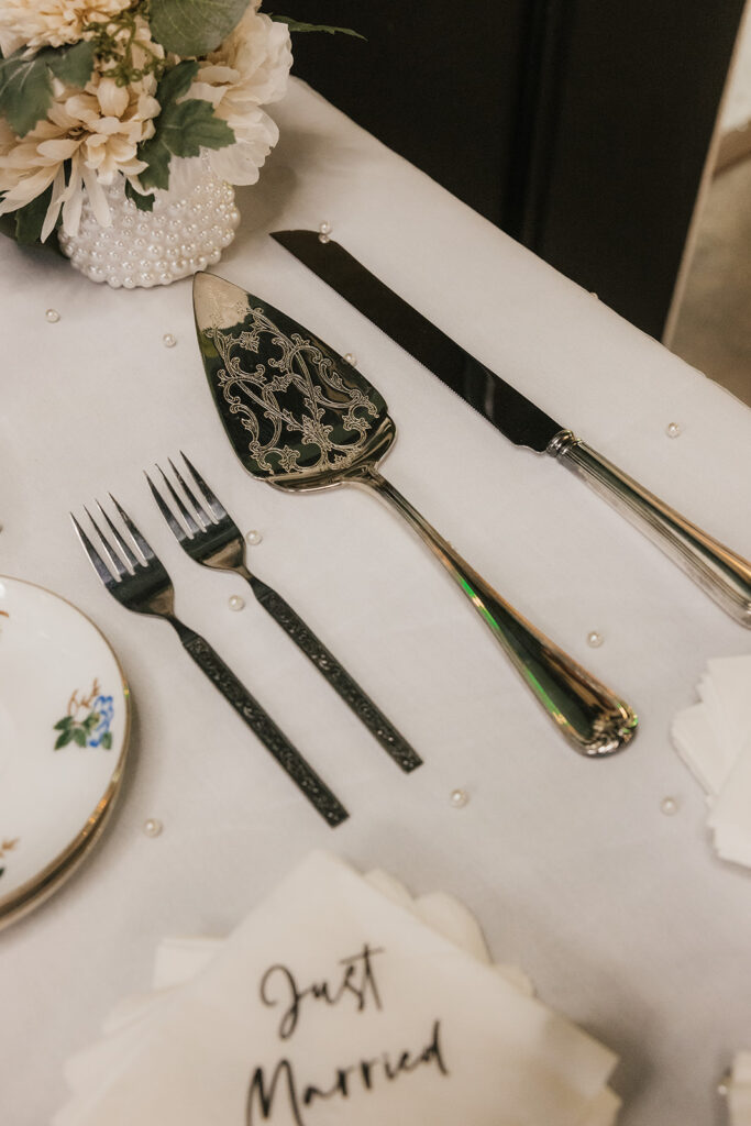 Close-up of a table setting with a cake server, knife, two forks, a plate with floral design, and a napkin that reads "Just Married" next to a white floral centerpiece.