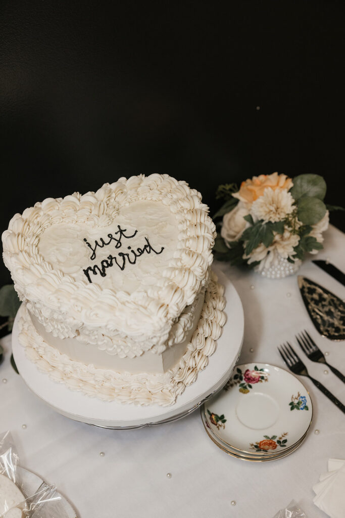 Close-up of a table setting with a cake server, knife, two forks, a plate with floral design, and a napkin that reads "Just Married" next to a white floral centerpiece.