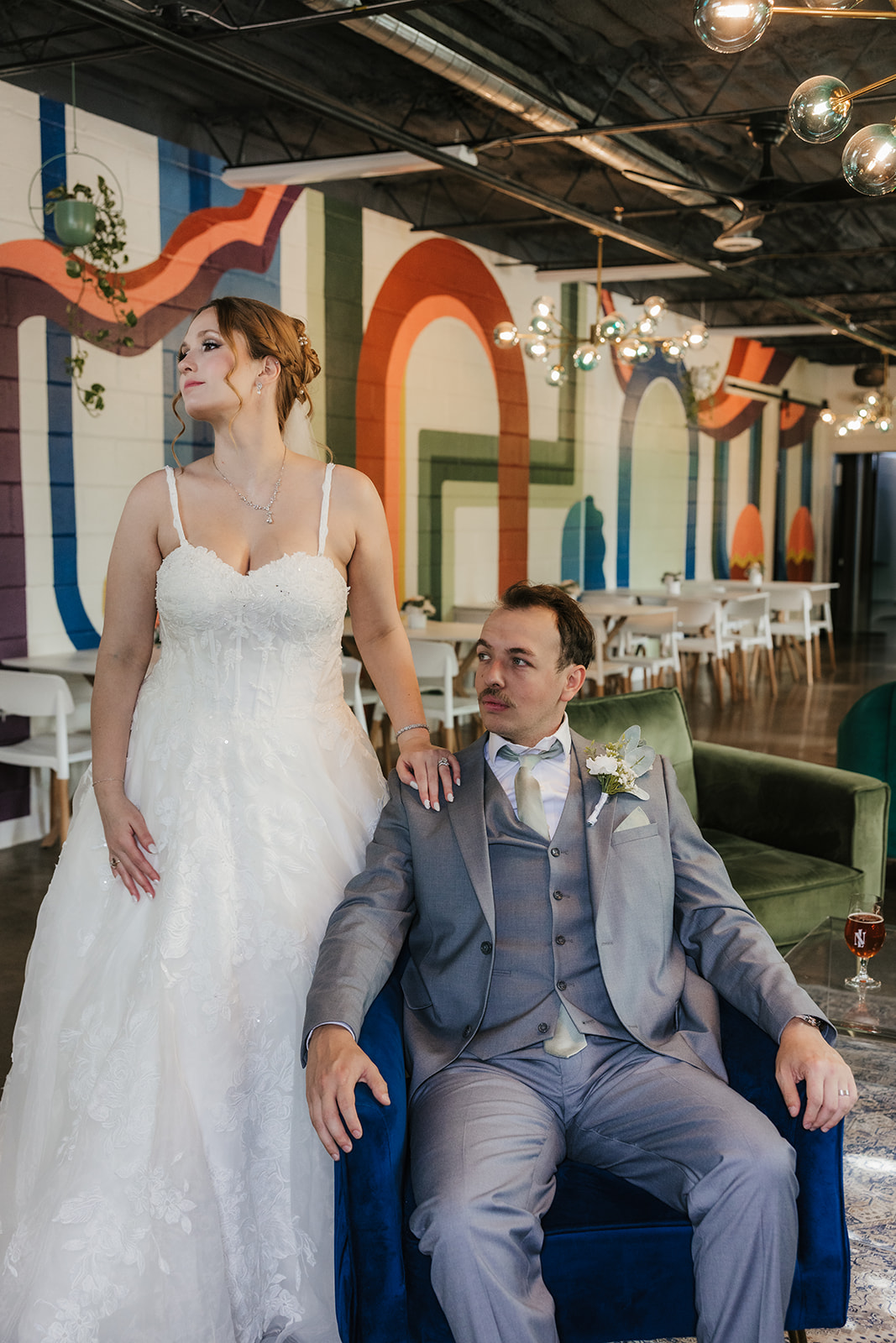 A bride in a white gown sits on a chair while a groom in a gray suit stands beside her in a decorated indoor venue A bride and groom share a dance at their wedding reception indoors, with guests and decorations visible in the background at a Camp Loughridge Wedding