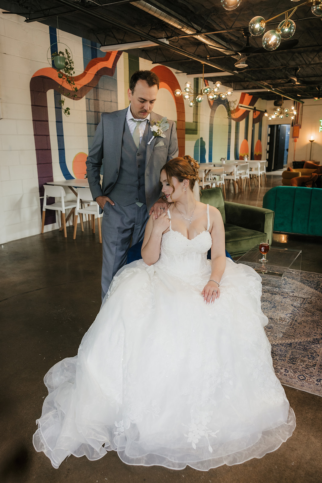 A bride in a white gown sits on a chair while a groom in a gray suit stands beside her in a decorated indoor venue A bride and groom share a dance at their wedding reception indoors, with guests and decorations visible in the background at a Camp Loughridge Wedding