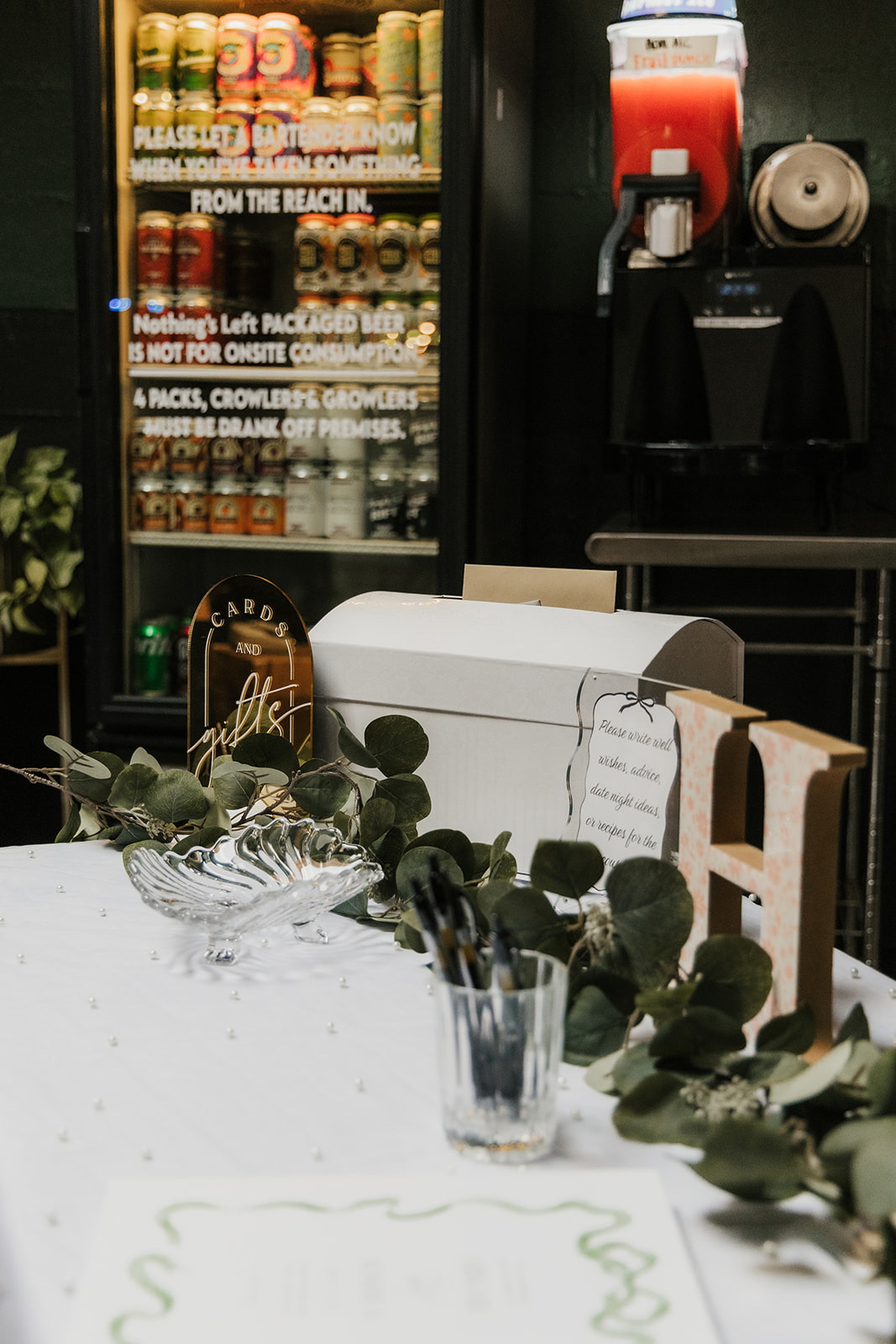 A modern restaurant interior with wooden tables, black cushioned benches, green plants, and pendant lights, with a person walking in the background at a Camp Loughridge Wedding