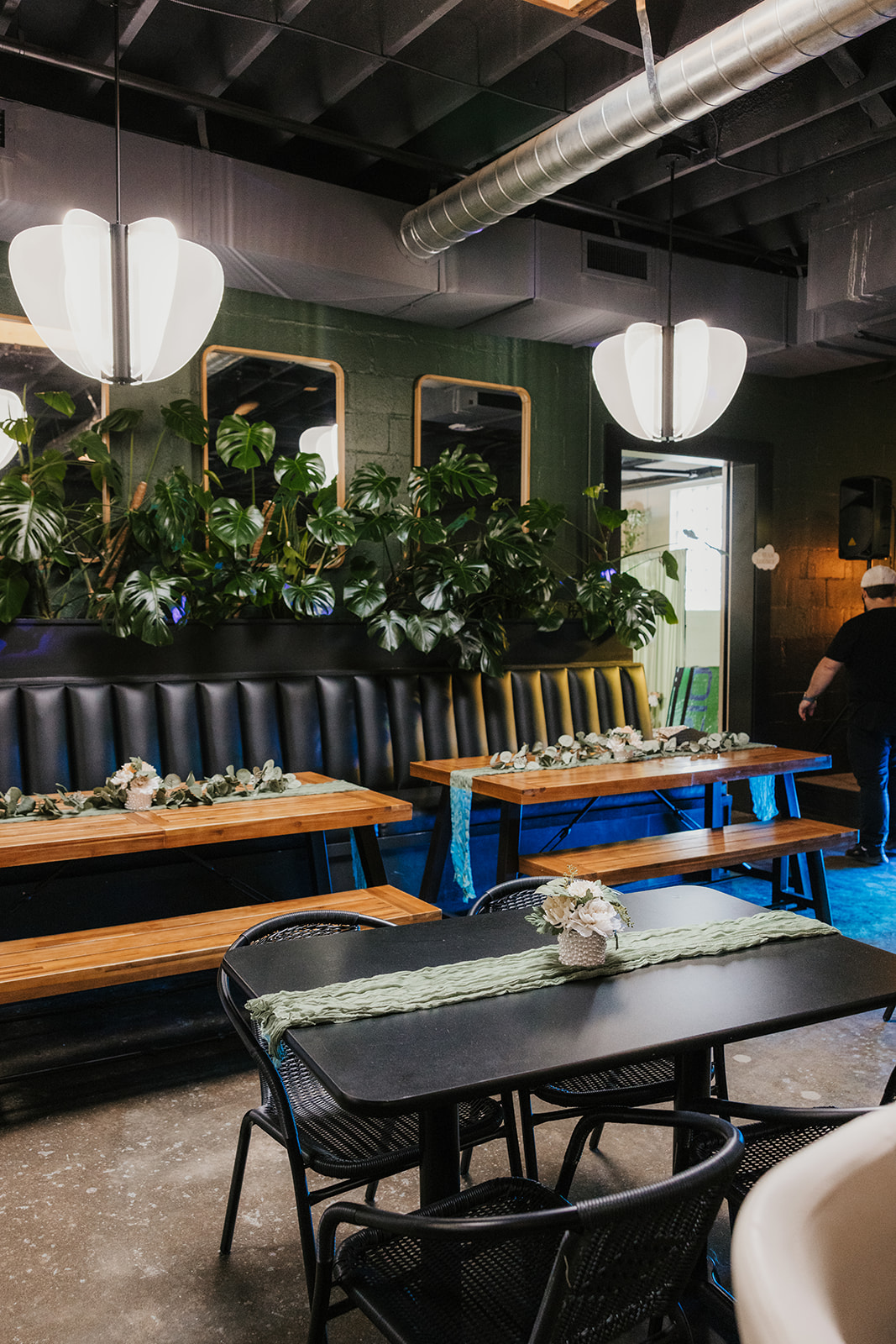 A modern restaurant interior with wooden tables, black cushioned benches, green plants, and pendant lights, with a person walking in the background at a Camp Loughridge Wedding