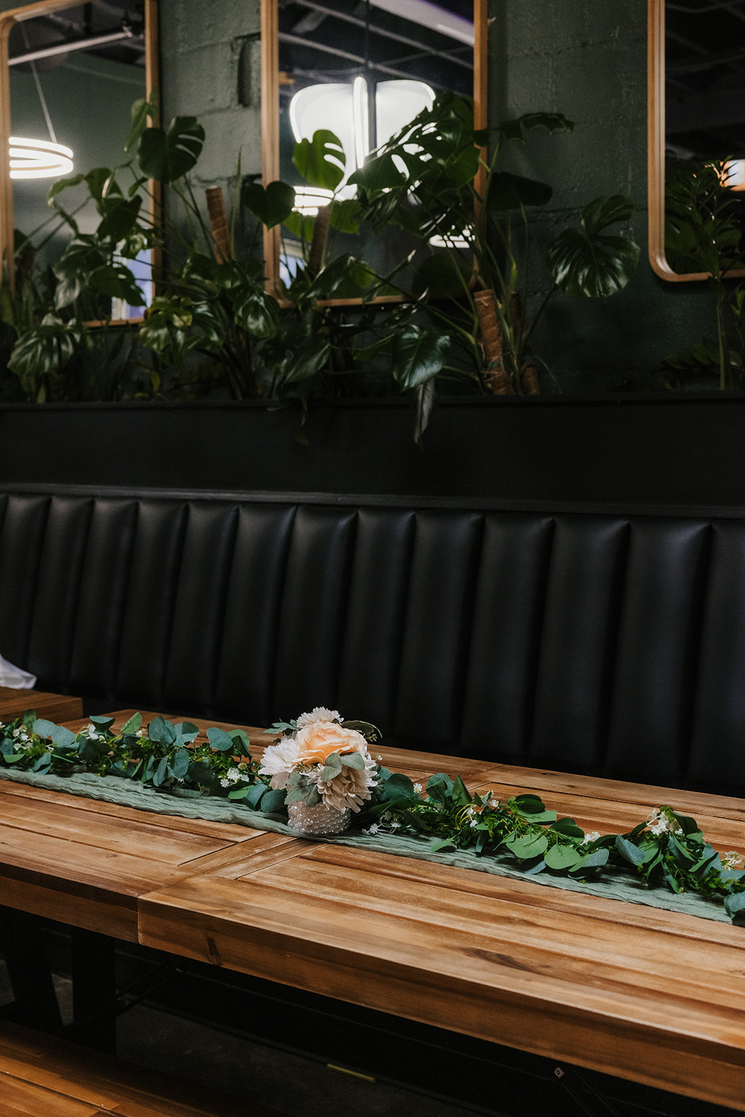 A modern restaurant interior with wooden tables, black cushioned benches, green plants, and pendant lights, with a person walking in the background at a Camp Loughridge Wedding