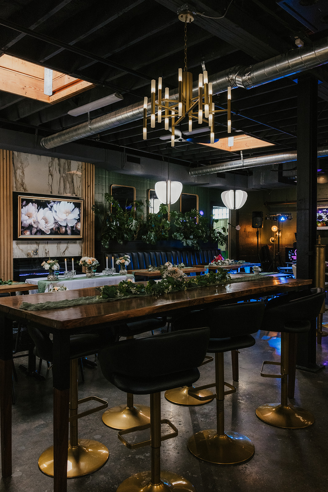A modern restaurant interior with wooden tables, black cushioned benches, green plants, and pendant lights, with a person walking in the background at a Camp Loughridge Wedding
