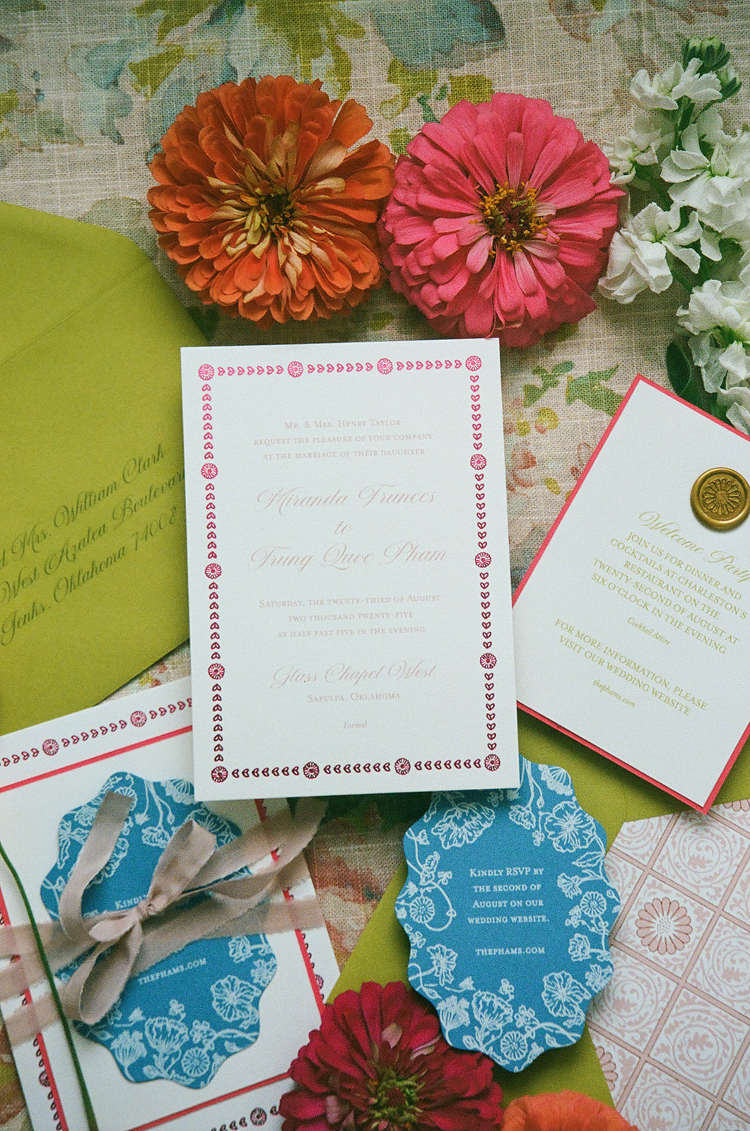 Flat lay of floral-themed stationery, place cards, a ribbon-tied invitation with a blue wax seal, yellow and orange flowers, and a green envelope on a floral fabric background. | unique wedding design
