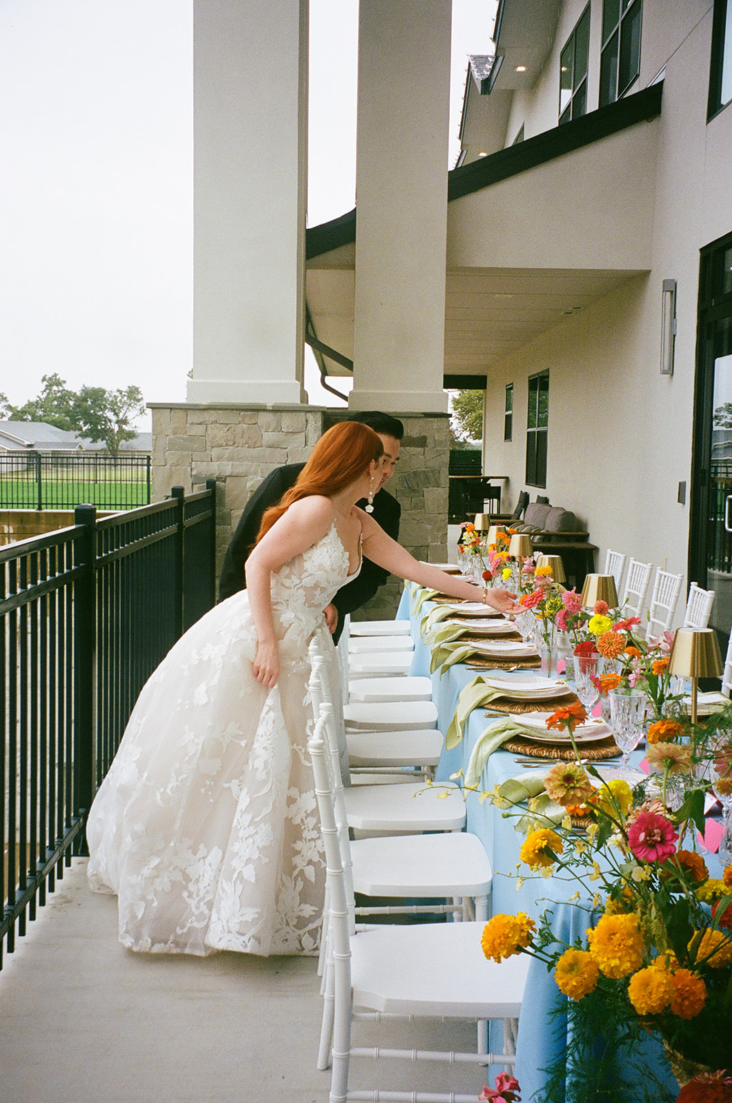 A bride and groom stand together by a decorated outdoor table set with plates, flowers, and candles, with a fence and trees in the background. | unique wedding design