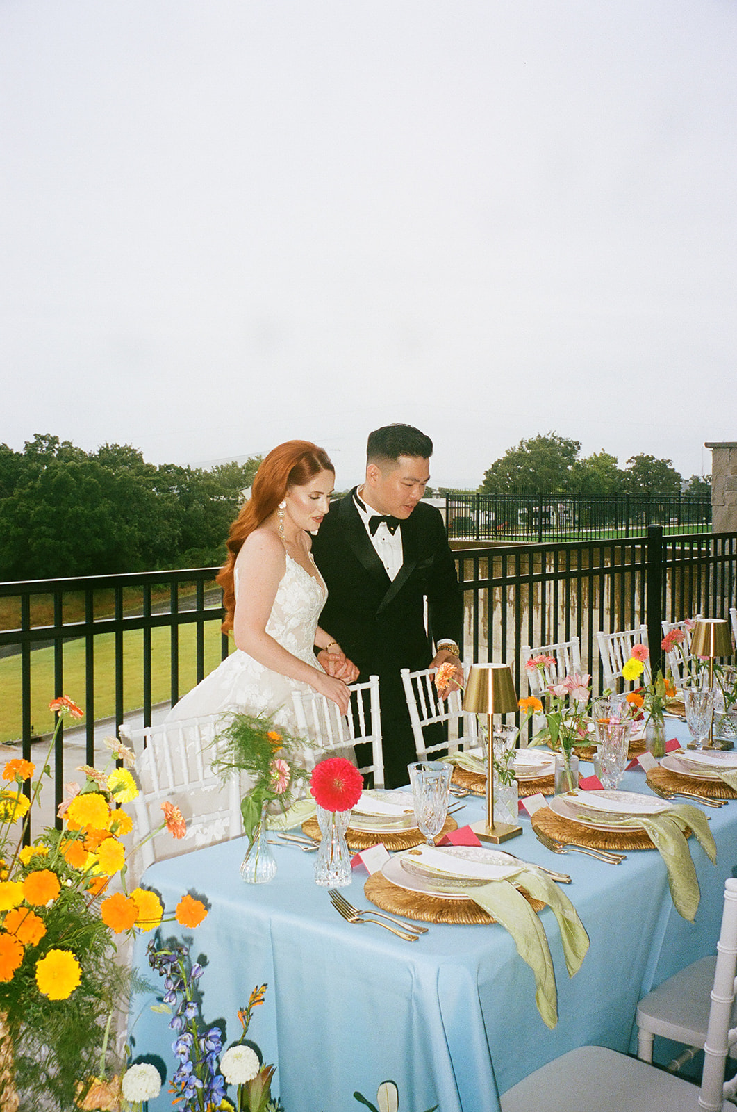A bride and groom stand together by a decorated outdoor table set with plates, flowers, and candles, with a fence and trees in the background. | unique wedding design