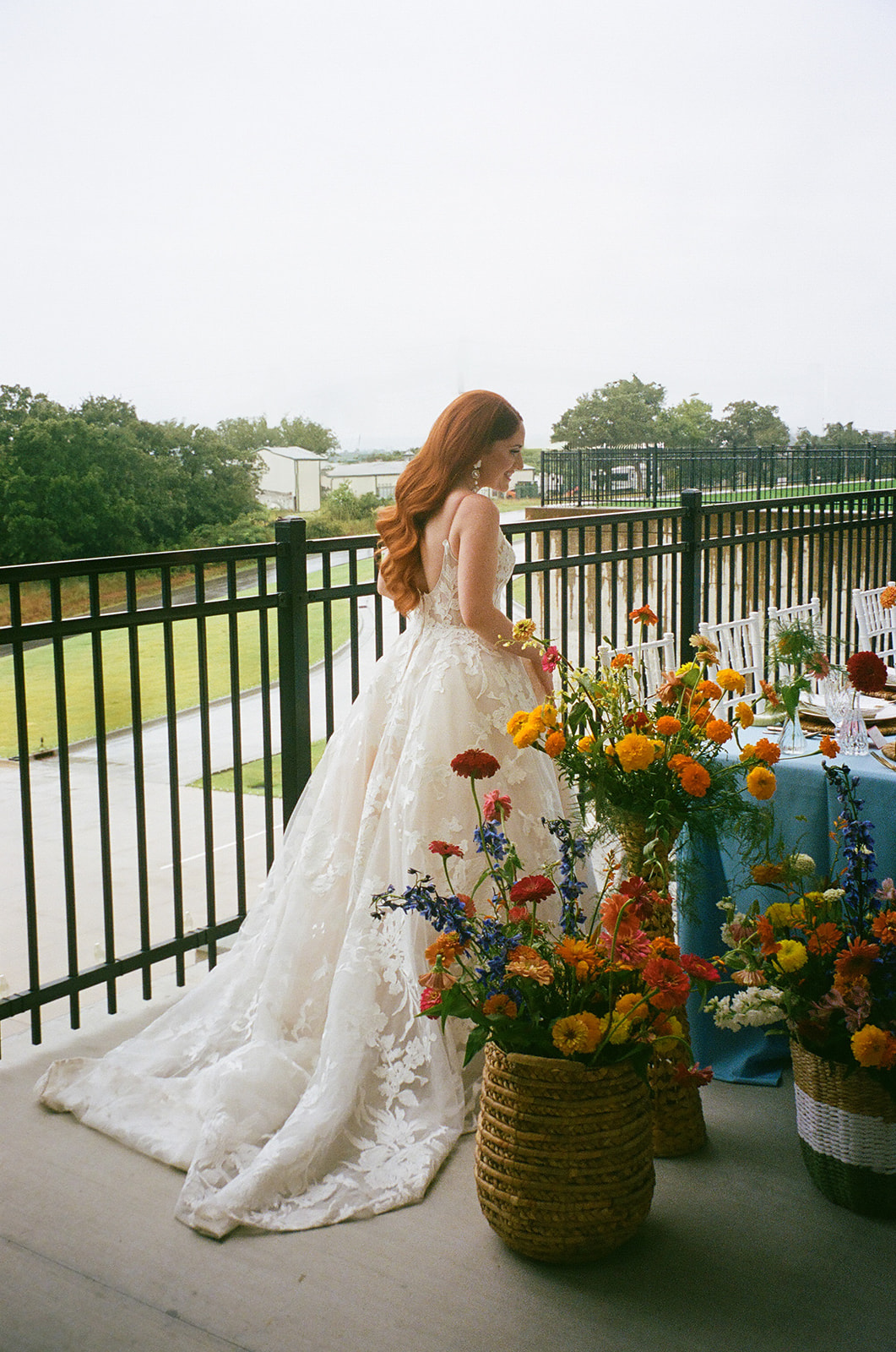 A bride and groom stand together by a decorated outdoor table set with plates, flowers, and candles, with a fence and trees in the background. | unique wedding design