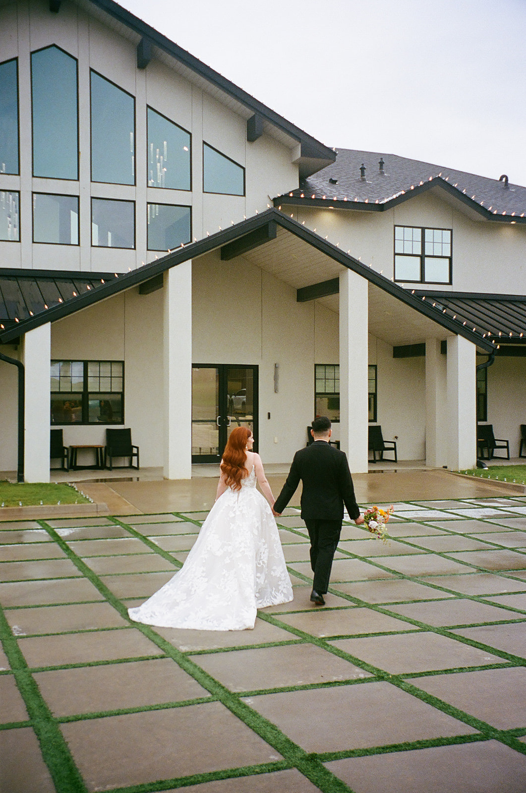 A bride in a white dress and a groom in a black suit walk hand in hand across a paved courtyard toward a modern white building | unique wedding design