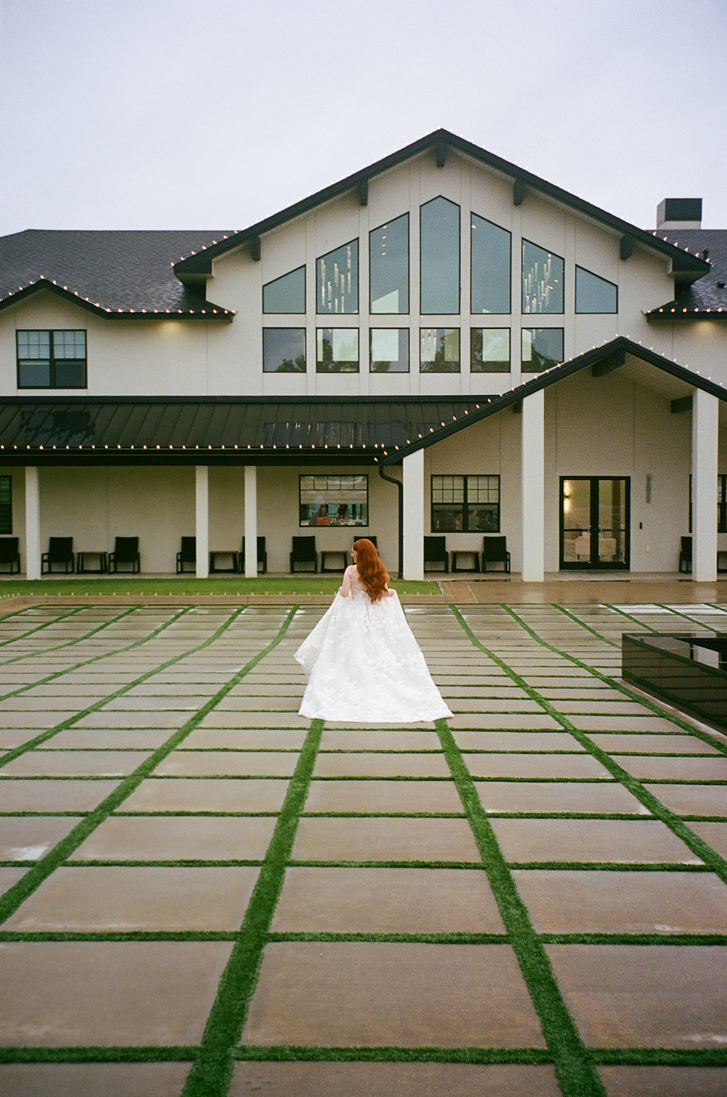 A bride in a white dress and a groom in a black suit walk hand in hand across a paved courtyard toward a modern white building | unique wedding design