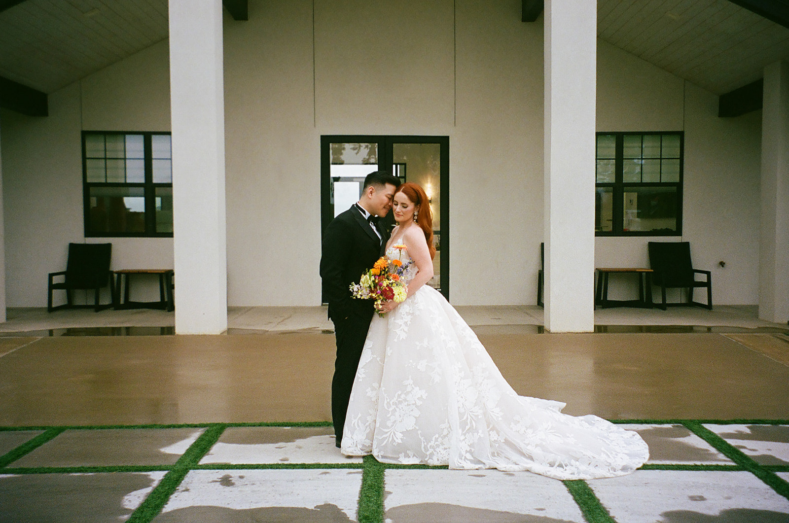 A bride in a white gown and a groom in a black suit stand together outside a modern building, with the groom kissing the bride's forehead.
