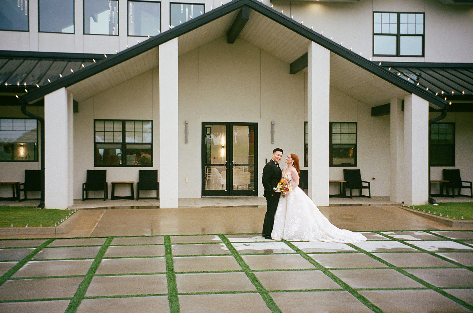A bride in a white gown and a groom in a black suit stand together outside a modern building, with the groom kissing the bride's forehead.
