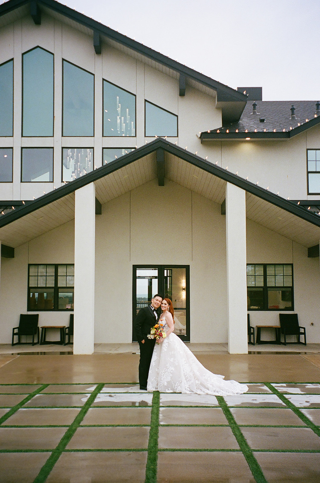 A bride in a white dress and a groom in a black suit walk hand in hand across a paved courtyard toward a modern white building | unique wedding design