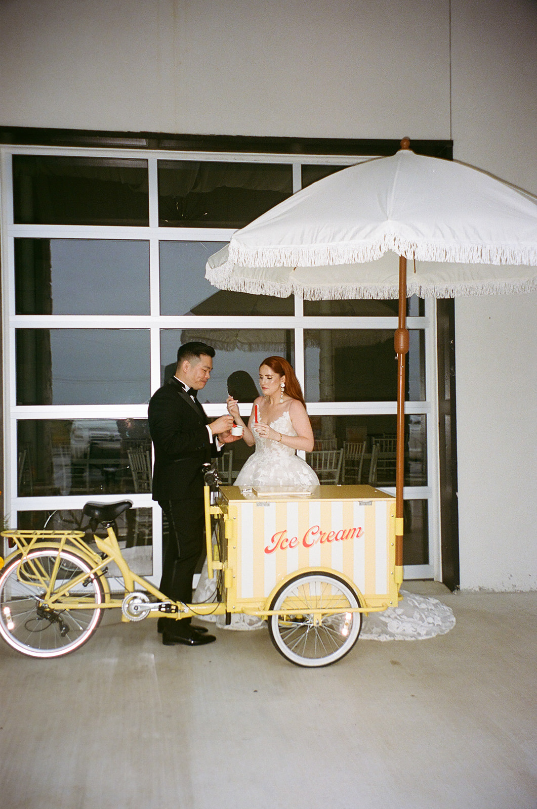 A bride in a white dress and a groom in a black suit walk hand in hand across a paved courtyard toward a modern white building | unique wedding design