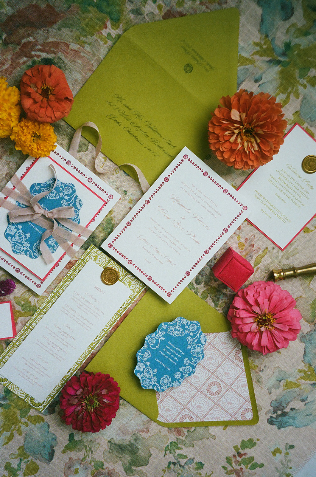 Flat lay of floral-themed stationery, place cards, a ribbon-tied invitation with a blue wax seal, yellow and orange flowers, and a green envelope on a floral fabric background. | unique wedding design