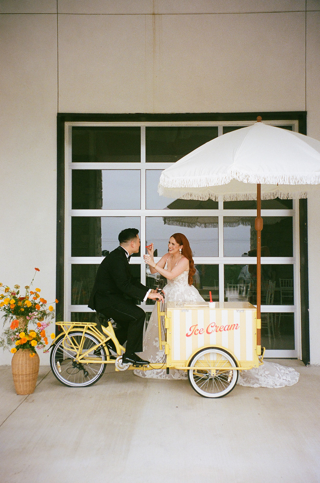 A bride in a white dress and a groom in a black suit walk hand in hand across a paved courtyard toward a modern white building | unique wedding design