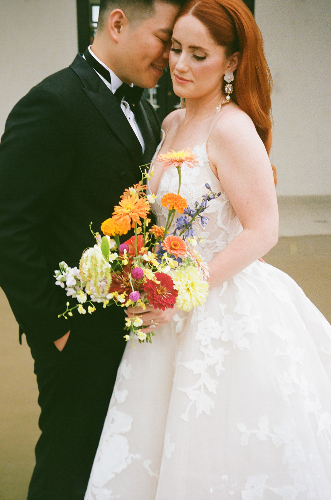 A bride in a white gown and a groom in a black suit stand together outside a modern building, with the groom kissing the bride's forehead.
