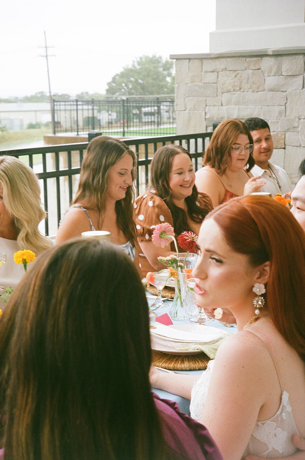 A group of people sit around a table outdoors, engaged in conversation, with plates, drinks, and flowers arranged on the table.