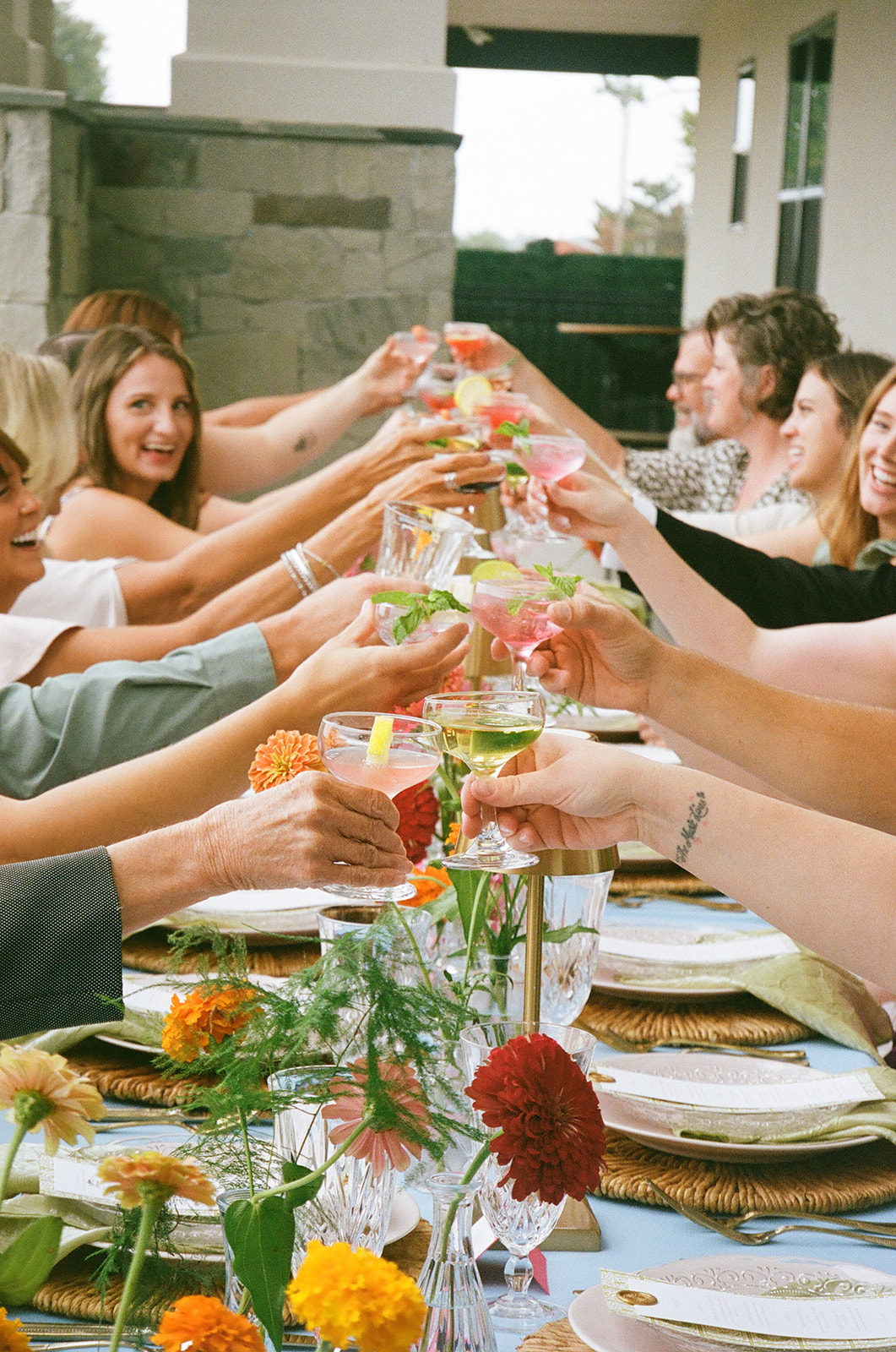 A group of people sit around a table outdoors, engaged in conversation, with plates, drinks, and flowers arranged on the table.