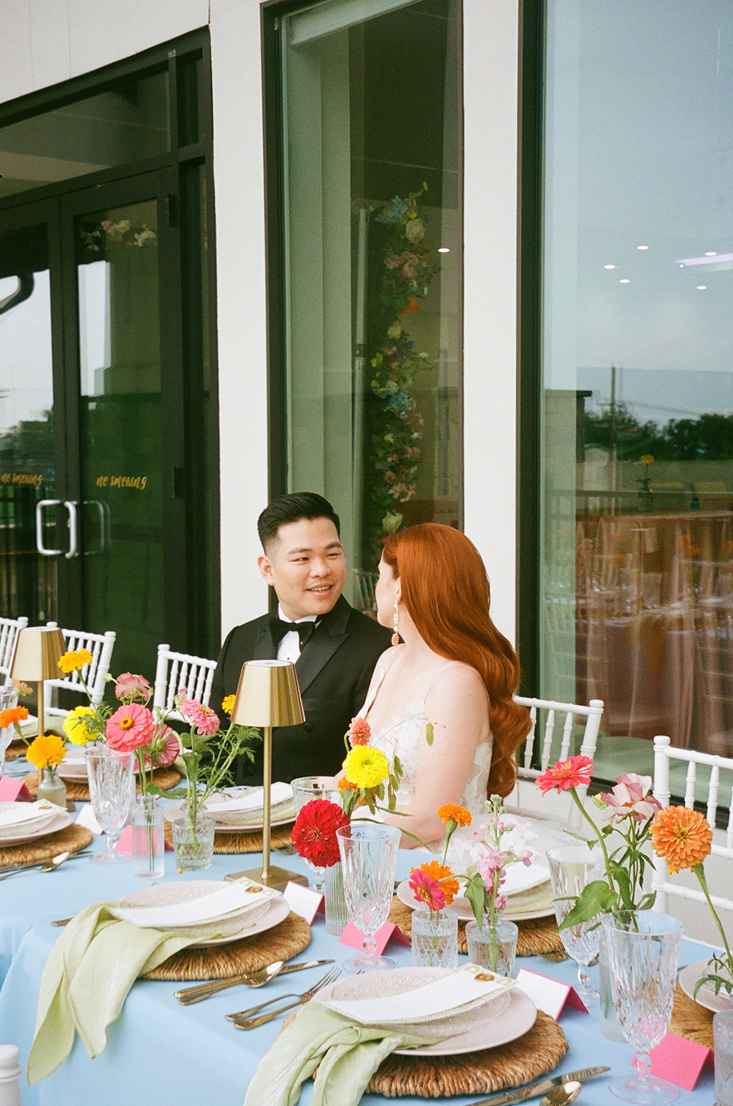 A group of people sit around a table outdoors, engaged in conversation, with plates, drinks, and flowers arranged on the table.