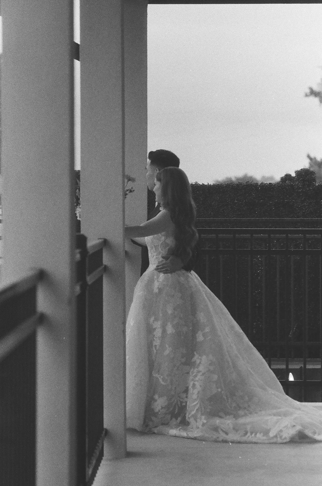 A couple dressed in formal attire, with the woman in a wedding gown, stands on a balcony looking outward in a black and white photo | unique wedding design