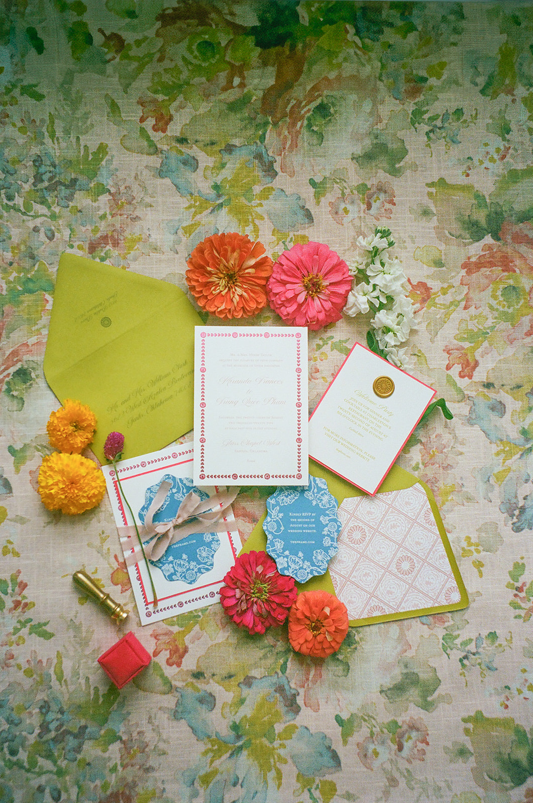 Flat lay of floral-themed stationery, place cards, a ribbon-tied invitation with a blue wax seal, yellow and orange flowers, and a green envelope on a floral fabric background. | unique wedding design
