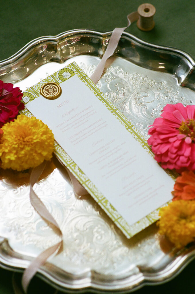Flat lay of floral-themed stationery, place cards, a ribbon-tied invitation with a blue wax seal, yellow and orange flowers, and a green envelope on a floral fabric background. | unique wedding design