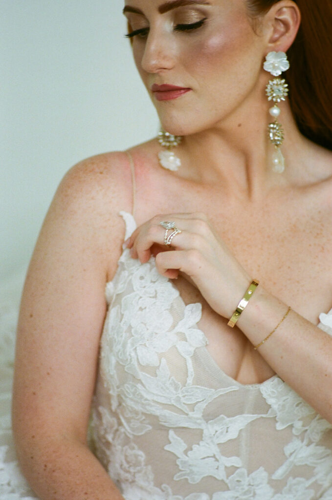 A woman with long red hair in loose waves wears a white, floral-patterned, sleeveless gown and stands near large windows, facing slightly away from the camera.