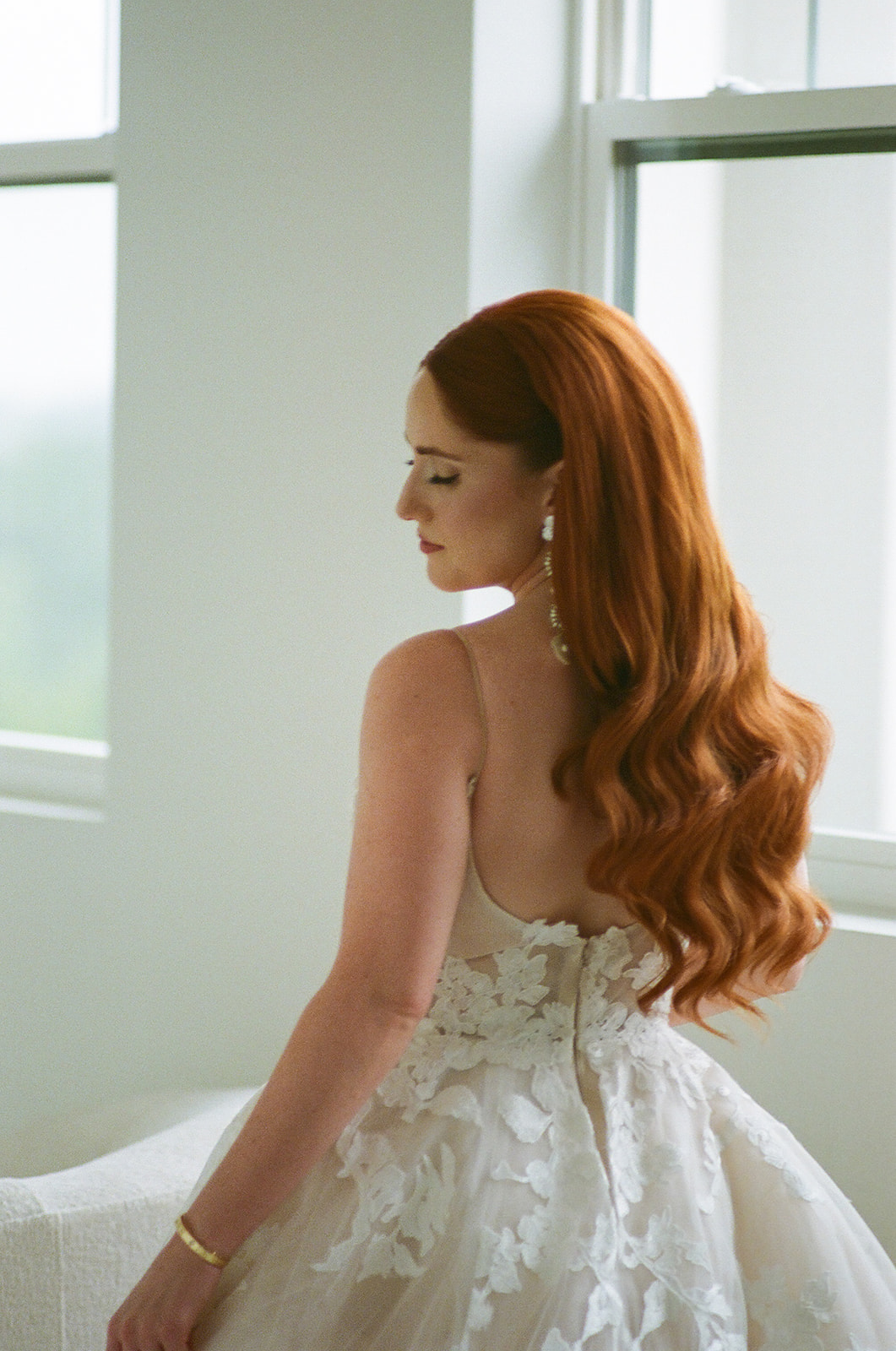 A woman with long red hair in loose waves wears a white, floral-patterned, sleeveless gown and stands near large windows, facing slightly away from the camera.
