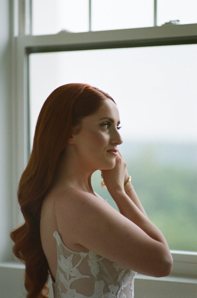 A woman with long red hair in loose waves wears a white, floral-patterned, sleeveless gown and stands near large windows, facing slightly away from the camera.