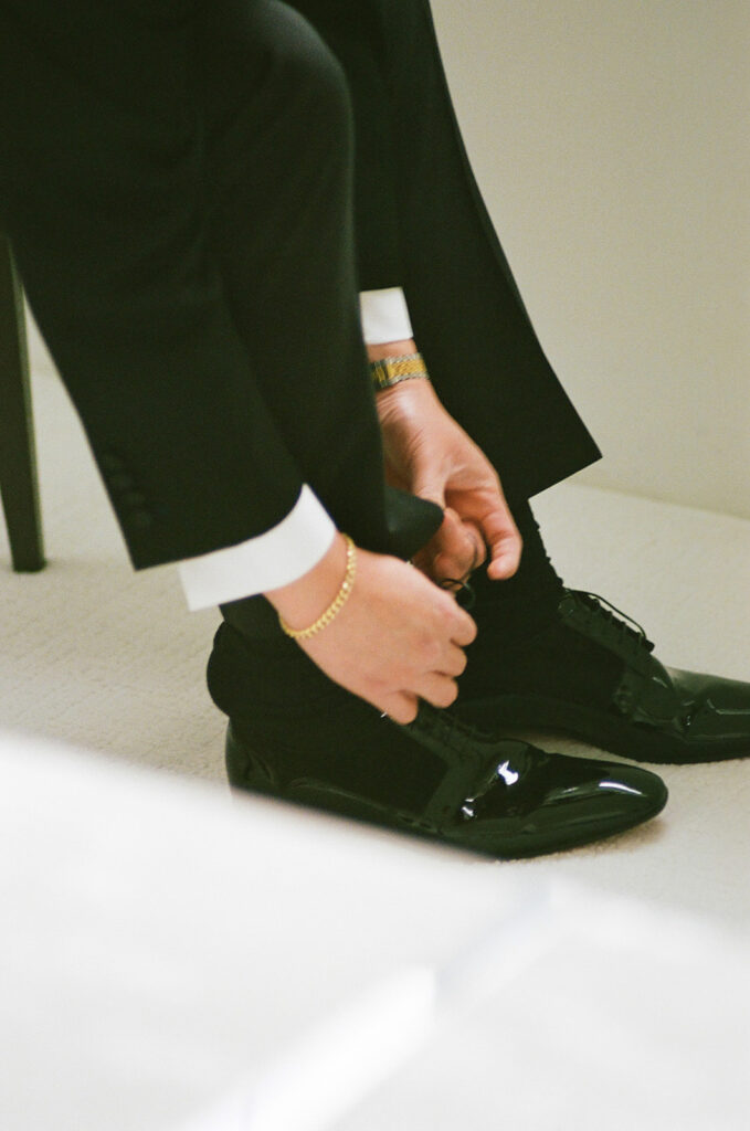 Person in a black suit and white shirt is sitting and tying the laces of shiny black dress shoes.