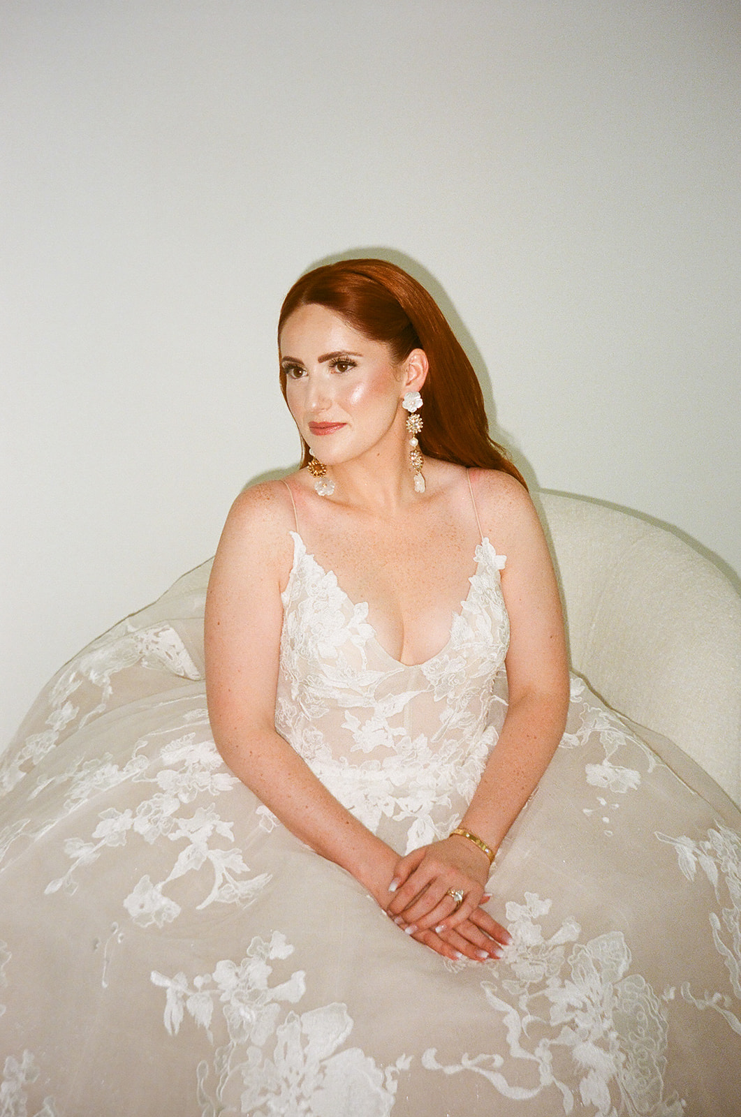 A woman with long red hair in loose waves wears a white, floral-patterned, sleeveless gown and stands near large windows, facing slightly away from the camera.