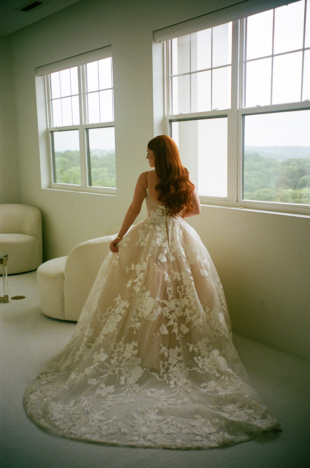 A woman with long red hair in loose waves wears a white, floral-patterned, sleeveless gown and stands near large windows, facing slightly away from the camera.