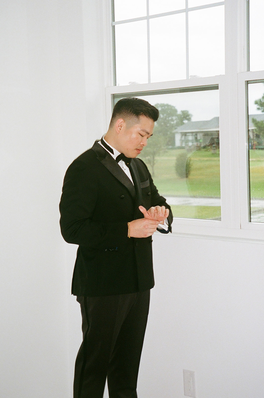 A man in a black tuxedo stands indoors by a window, looking down at his wristwatch.