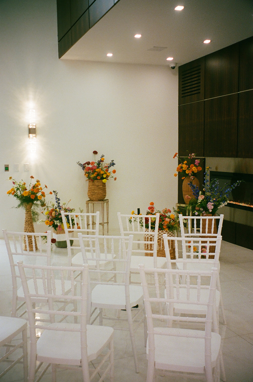 A small indoor event space with white chairs arranged in rows, floral arrangements in vases, and a modern fireplace against a wood-paneled wall.