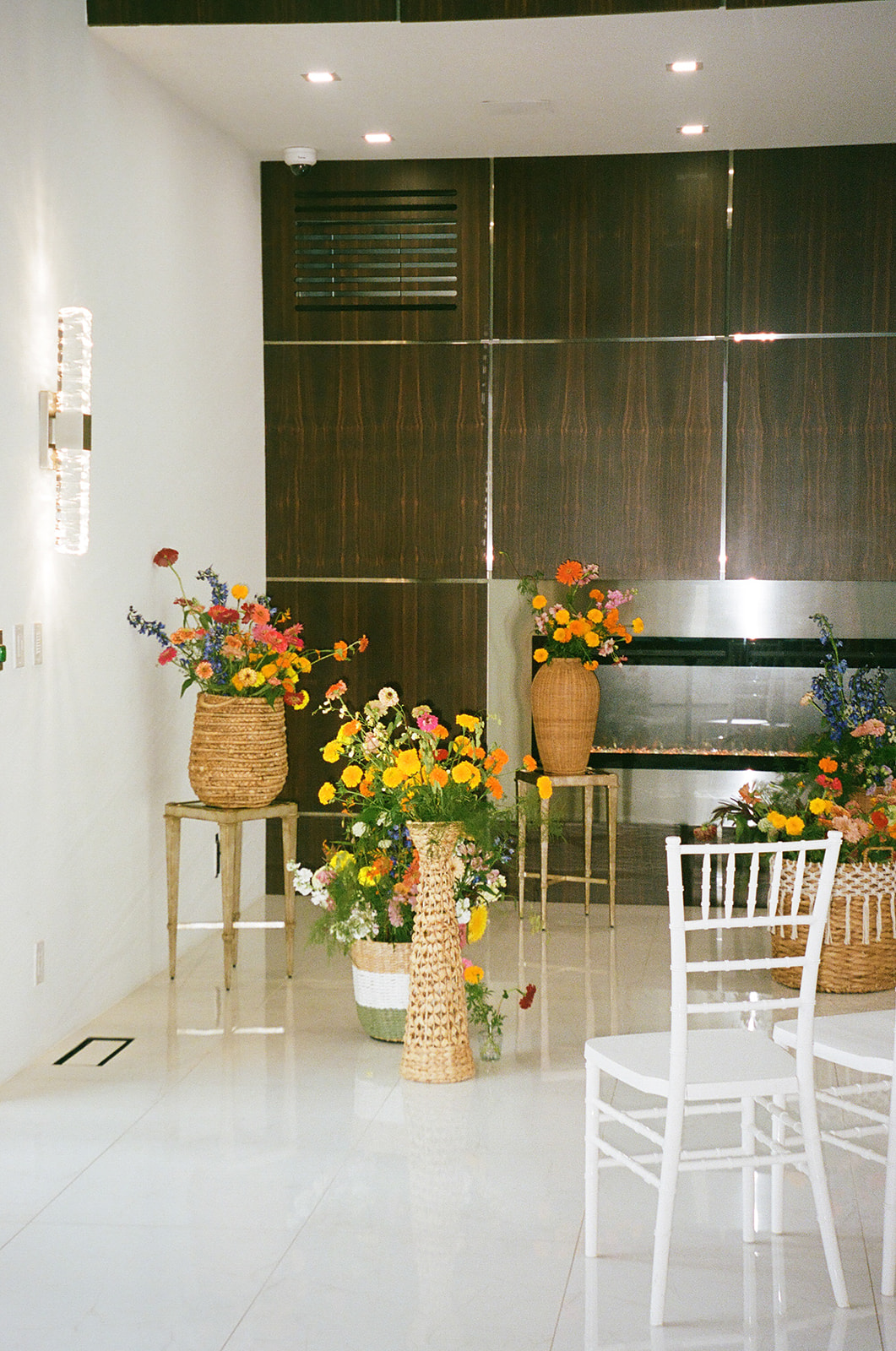 A small indoor event space with white chairs arranged in rows, floral arrangements in vases, and a modern fireplace against a wood-paneled wall.