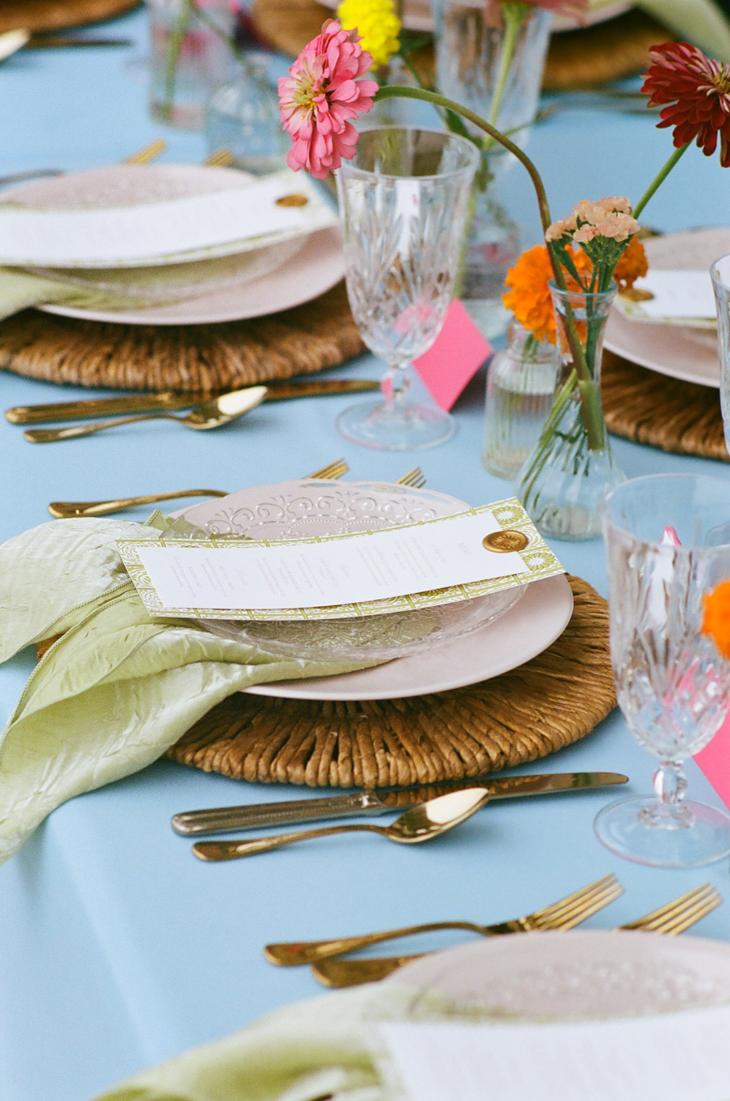 A long dining table with a light blue tablecloth, gold cups, glassware, and colorful flower arrangements is set outdoors beside a stone wall.