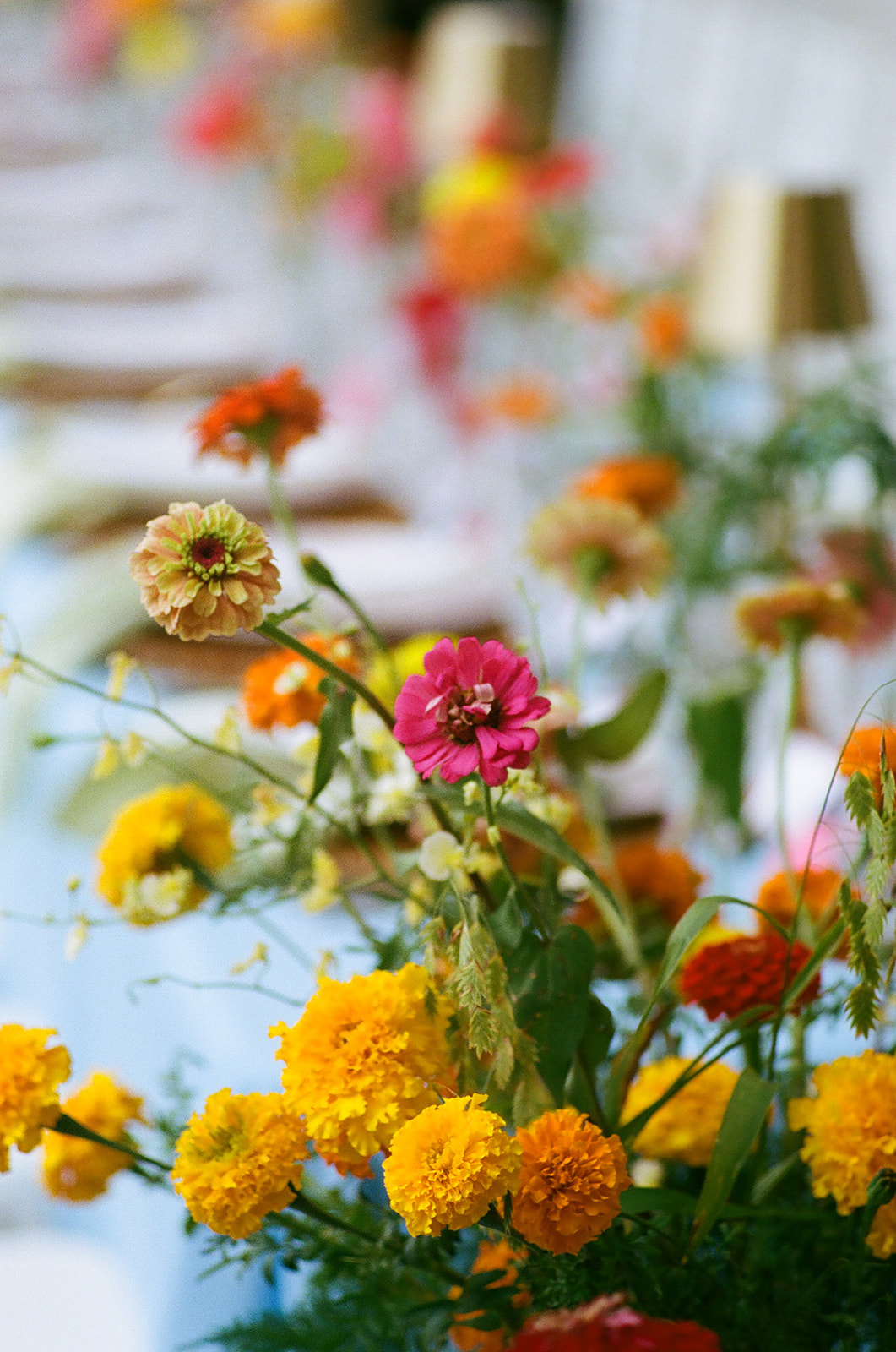 A long dining table with a light blue tablecloth, gold cups, glassware, and colorful flower arrangements is set outdoors beside a stone wall.