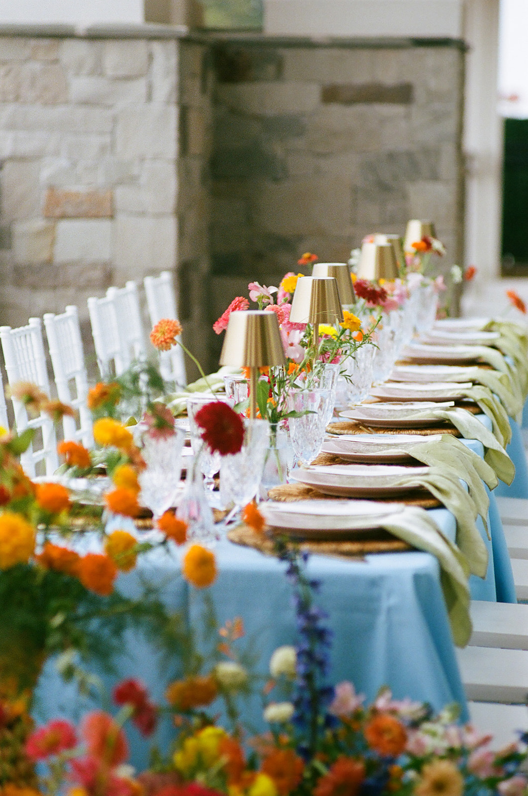 A long dining table with a light blue tablecloth, gold cups, glassware, and colorful flower arrangements is set outdoors beside a stone wall.