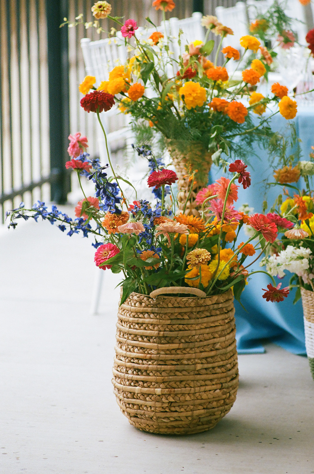 A long dining table with a light blue tablecloth, gold cups, glassware, and colorful flower arrangements is set outdoors beside a stone wall.