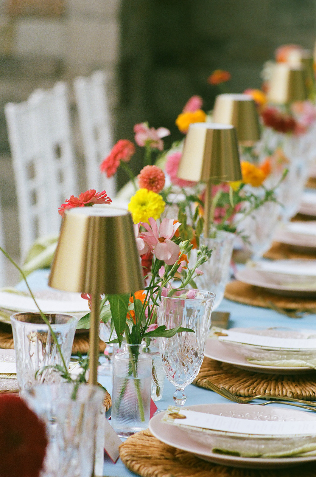A long dining table with a light blue tablecloth, gold cups, glassware, and colorful flower arrangements is set outdoors beside a stone wall.